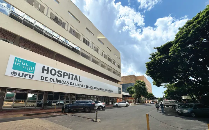 Entrada principal do Hospital de Clínicas da UFU, em Uberlândia, com fachada identificada e árvores ao lado, em fotografia de 2024