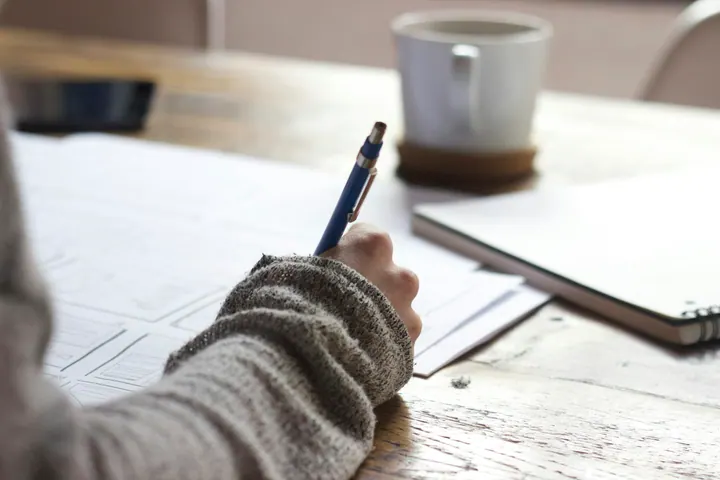 Pessoa fazendo anotações em papel, com caderno e xícara sobre uma mesa