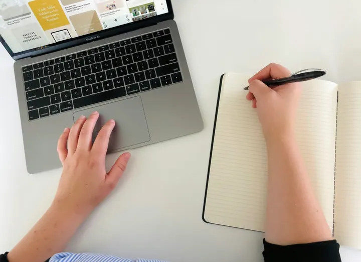 Vista superior de mãos sobre um notebook e um caderno, com uma caneta pronta para anotar