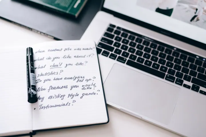 Caderno pautado com perguntas manuscritas e marcador preto ao lado do teclado de um computador